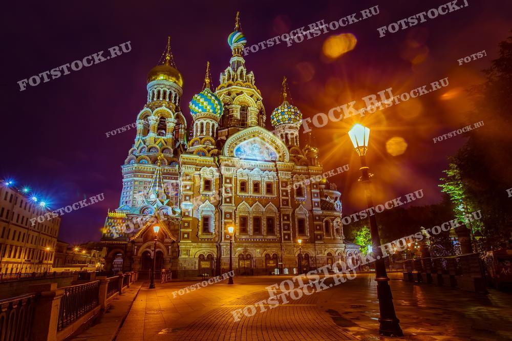 Фотография демонстрирует Спас на Крови в Санкт-Петербурге, одну из самых знаковых достопримечательностей города, освещённую вечерними огнями. Свет фонарей добавляет теплоты и таинственности кадру, подчеркивая красочные купола и богатое убранство собора. Это изображение подойдёт для использования в туристических гидах, культурно-исторических обзорах, а также как элемент декора в гостиницах или общественных пространствах, демонстрирующий красоту российской архитектуры.
